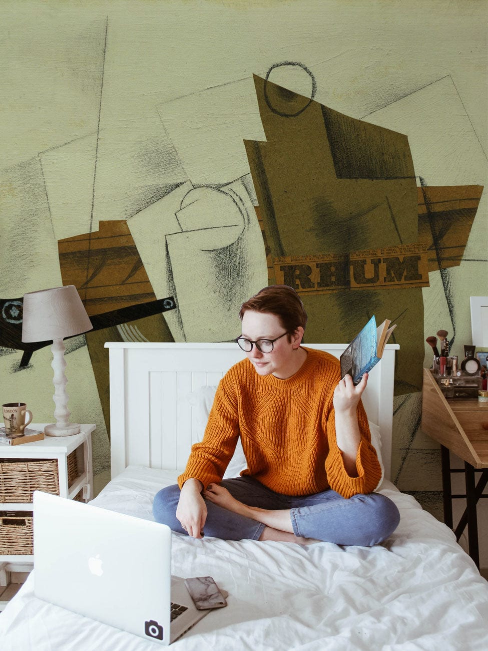 Personne lisant un livre sur un lit blanc avec un papier peint mural représentant une pipe, un verre et une bouteille de rhum