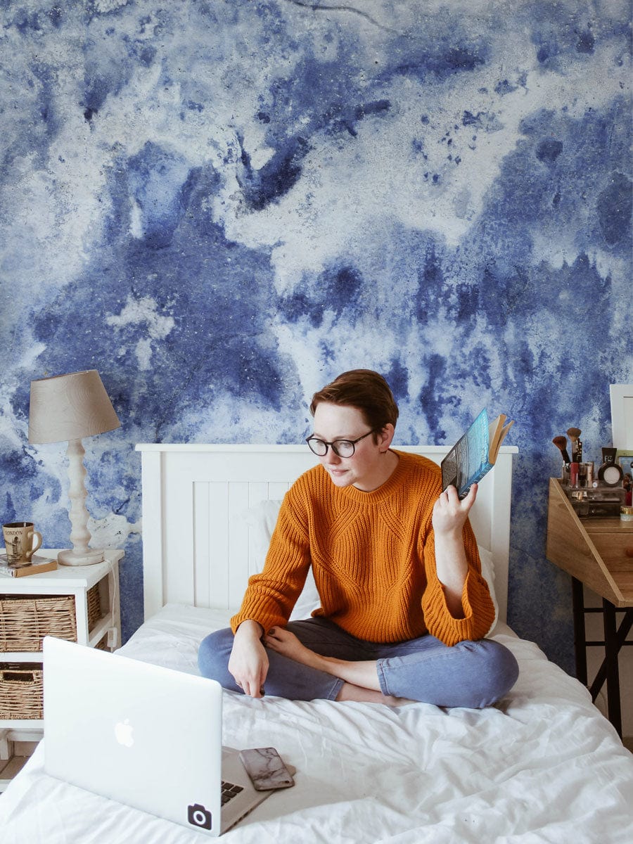 Person assise sur un lit avec un ordinateur et un livre, dans un décor mural tempête bleue